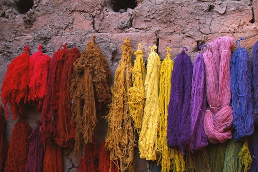 Various strands of brightly colored yarn or threads hang against a textured, earthy wall. The colors range from deep reds and browns to yellows, purples, pinks, and blues. The yarns are arranged in a way that contrasts with the rustic appearance of the background, suggesting a traditional or handmade context.