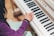A child wearing a purple sweater is playing a Yamaha piano. The child's left hand is visible, pressing down on a key. There is a small sticker on the child's wrist.