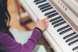 A child wearing a purple sweater is playing a Yamaha piano. The child's left hand is visible, pressing down on a key. There is a small sticker on the child's wrist.