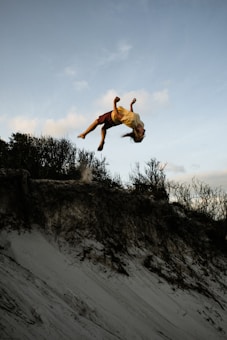 A person is performing a mid-air flip above a sandy slope with sparse vegetation in the background. The sky appears clear with a few clouds, indicating a calm weather setting. The person's hair and clothing reflect motion, emphasizing the dynamic action of the flip.