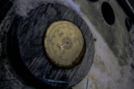 A vintage barometer mounted on a wooden piece with an aged and weathered appearance. The barometer has a circular dial with various readings like rain, change, and very dry. The background consists of a rough surface and some snow or frost around the edges.