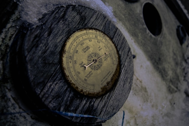 A vintage barometer mounted on a wooden piece with an aged and weathered appearance. The barometer has a circular dial with various readings like rain, change, and very dry. The background consists of a rough surface and some snow or frost around the edges.