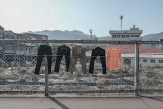 Clothing items hang to dry on a wire fence under a clear sky, with buildings and mountains in the background. The clothes include dark and light-colored pants and an orange shirt. The setting appears to be an industrial or urban area with vegetation growing in the foreground.