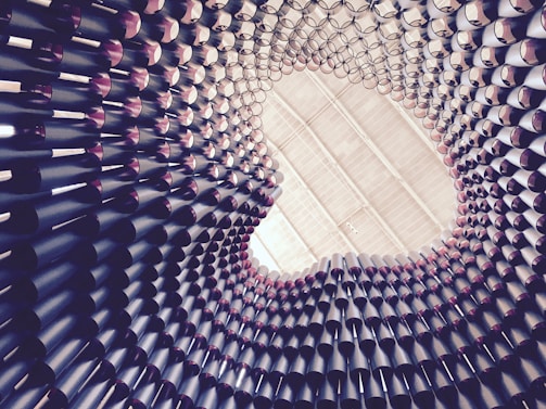A cylindrical structure made up of numerous stacked and layered tubes creates a spiraling tunnel-like visual. The tubes are arranged in a pattern that appears to extend upwards towards a circular opening at the top. The perspective suggests looking up from the inside of the structure, with the tubes casting shadows and creating a dynamic play of light and form.
