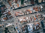 An aerial view of an urban area revealing densely packed buildings with varied rooftops in shades of rust and grey. The image includes multiple intersecting roads with visible traffic, comprised of cars and buses. The scene captures the complexity and busyness of city life.