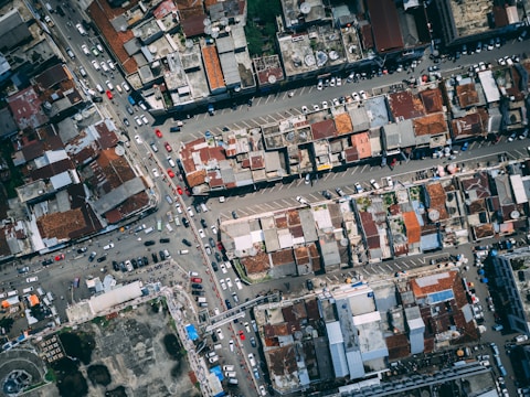 An aerial view of an urban area revealing densely packed buildings with varied rooftops in shades of rust and grey. The image includes multiple intersecting roads with visible traffic, comprised of cars and buses. The scene captures the complexity and busyness of city life.