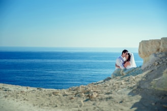 A couple embraces near the edge of a rocky cliff with the vast, calm blue ocean in the background. The woman is wearing a white wedding dress and veil, while the man is dressed in a light-colored shirt. The scene is serene and romantic, with clear skies above and sunlight illuminating the landscape.