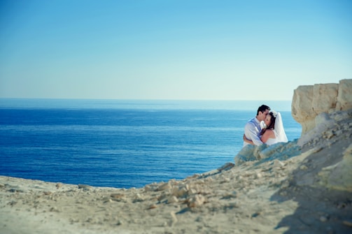 A couple embraces near the edge of a rocky cliff with the vast, calm blue ocean in the background. The woman is wearing a white wedding dress and veil, while the man is dressed in a light-colored shirt. The scene is serene and romantic, with clear skies above and sunlight illuminating the landscape.