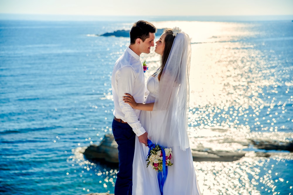 woman in wedding gown about to kiss man in white dress shirt while holding bouquet of flower near body of water at daytime