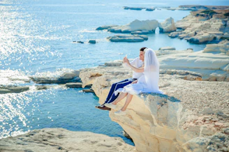 woman wearing wedding dress sitting beside man wearing white dress shirt on the stone tip facing the sea