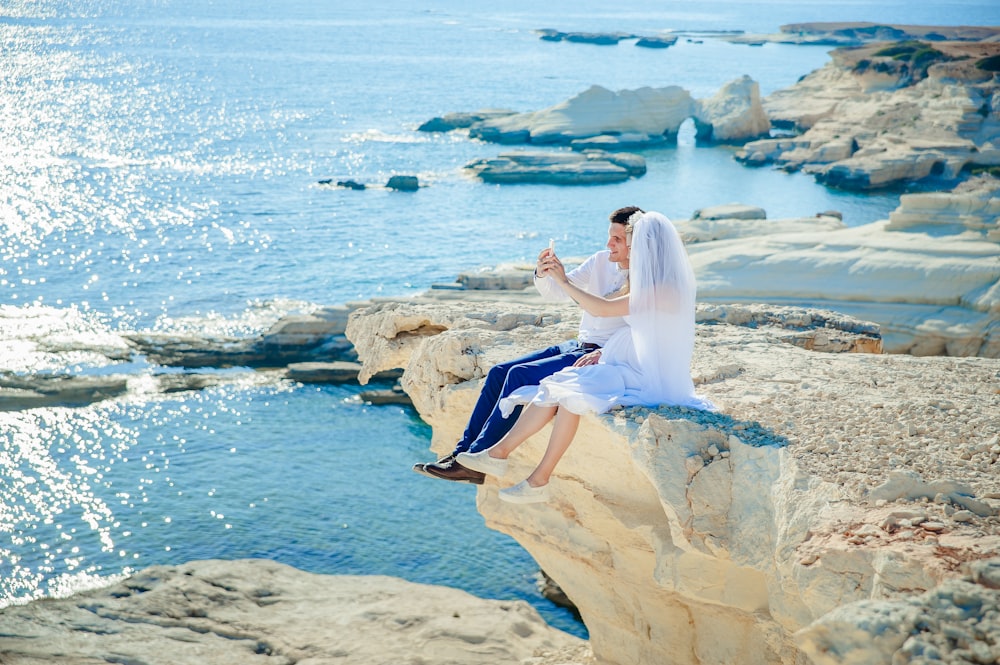 웨딩 드레스를 입은 여자가 바다를 향한 돌 끝에 흰 드레스 셔츠를 입은 남자 옆에 앉아 있습니다.