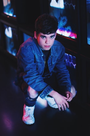 A young person crouches on the floor, dressed in a denim jacket and ripped jeans. The environment is dimly lit, with neon lights casting a blue and pink hue. The person has short, dark hair and is wearing white sneakers. They are positioned near screens or reflective surfaces that contribute to the glowing ambiance.