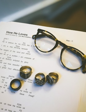 Sheet music for the song 'How He Loves' by John Mark McMillan is displayed on a surface, accompanied by a pair of tortoiseshell glasses and five rings of various designs. The rings have ornate patterns and distinct styles, positioned on the music sheet.