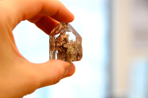 A hand holds a clear, faceted crystal with brown inclusions. The background is softly blurred, highlighting the delicate details of the crystal's inner textures.