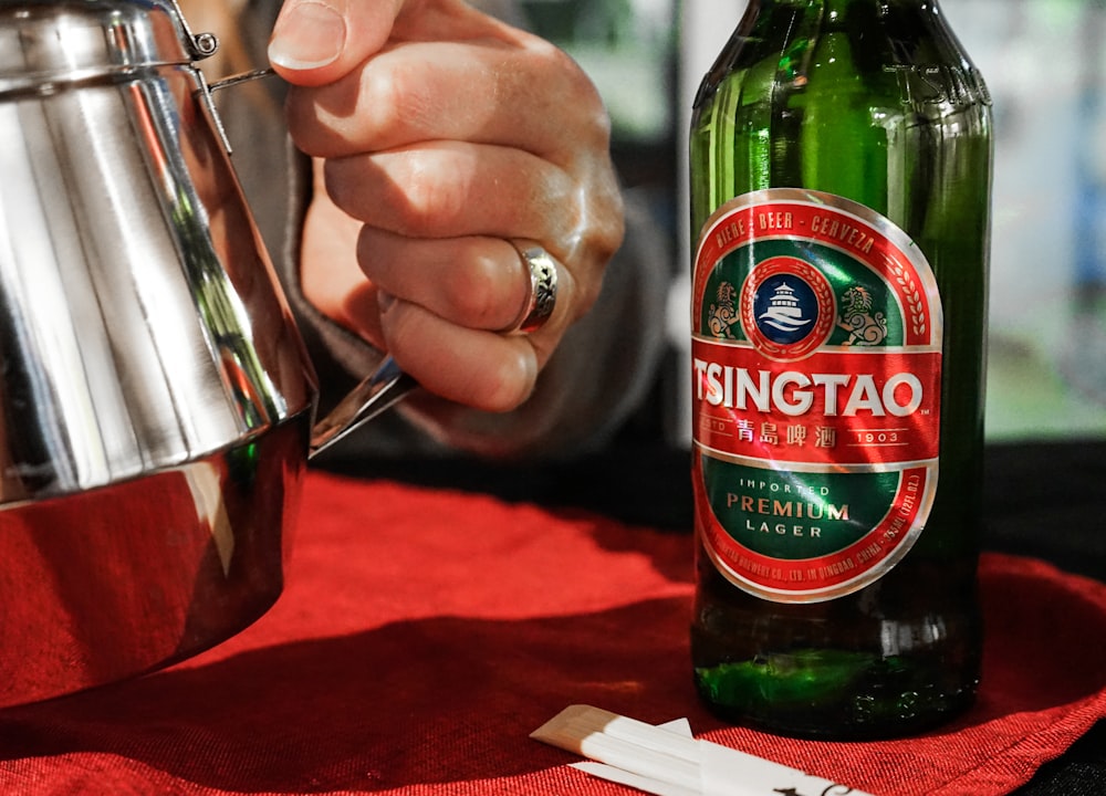 person holding silver pitcher beside a green Tsingtao bottle