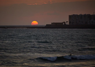 A vibrant sunset over a volcanic beach with crashing ocean waves and a silhouette of a surfer.