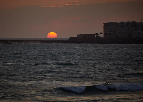 A vibrant sunset over a volcanic beach with crashing ocean waves and a silhouette of a surfer.