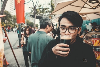A young man with glasses holds a glass of dark beverage to his mouth. He stands in an outdoor setting with several people around him, including a woman and an older man facing away. A street market stall is visible in the background, with umbrellas shading the area. A red flag with a yellow star hangs on a pole.