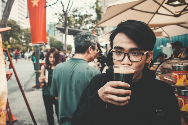 A young man with glasses holds a glass of dark beverage to his mouth. He stands in an outdoor setting with several people around him, including a woman and an older man facing away. A street market stall is visible in the background, with umbrellas shading the area. A red flag with a yellow star hangs on a pole.