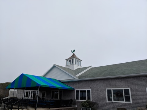 A building with a cupola on its roof is topped by a weather vane featuring a green figure of a lobster. The structure has a gray shingled facade and a blue and green striped awning over a porch area. The sky is overcast, creating a muted backdrop for the scene.