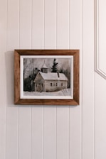 A framed painting depicts a rustic, snow-covered cabin surrounded by bare trees, placed against a wall with vertical panels.