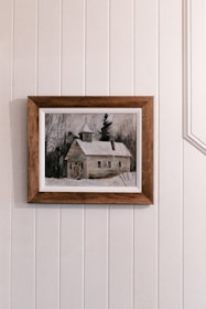 A framed painting depicts a rustic, snow-covered cabin surrounded by bare trees, placed against a wall with vertical panels.