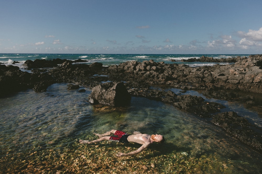 man in black and red shorts floating on shallow water surrounded by grey rocks during daytime