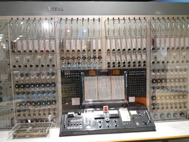 An intricate control panel featuring numerous dials, switches, and knobs arranged neatly on a complex machine. The panel includes various gauges and labels, with a vintage design suggesting it's an early computing or control system. The name 'TESLA' is engraved on the top.