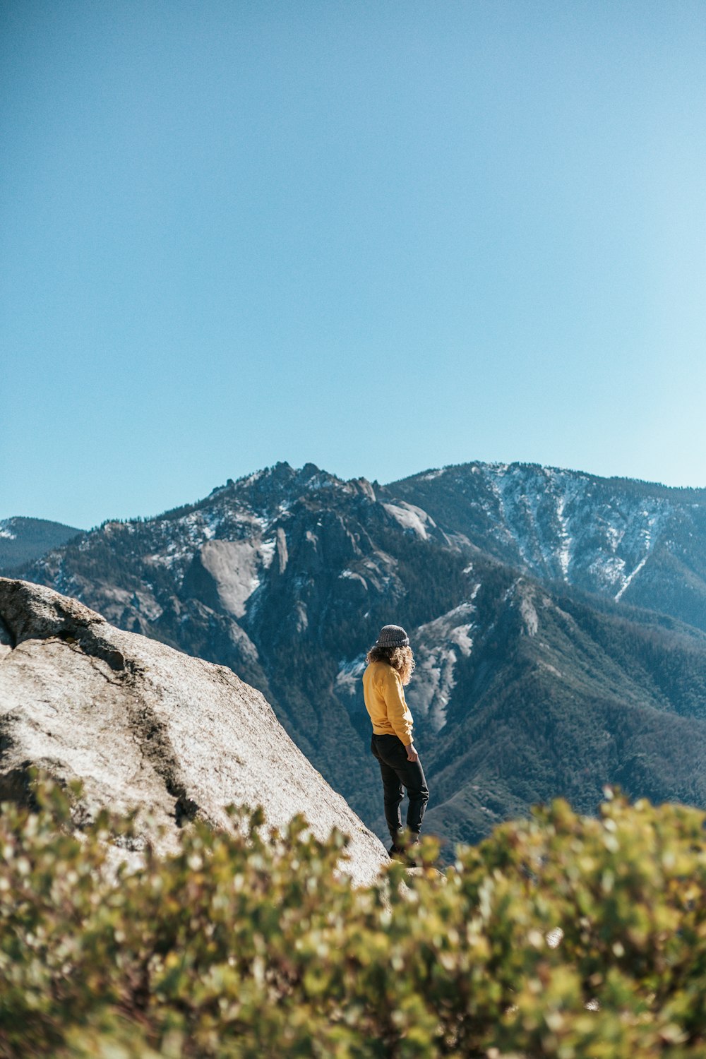 woman in yellow sweatshirt and black pants standing near cliff under blue sky during daytime