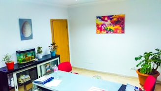 A well-lit office space features a table with documents, a water bottle, and a pen on it. There is a cabinet with glass doors, holding files and decorative items. On top of the cabinet, there is a small fish tank and two potted plants. The walls are adorned with two pieces of art: one depicting a natural scene with vibrant colors and another displaying a close-up of an egg. A large potted plant is situated near the right wall.