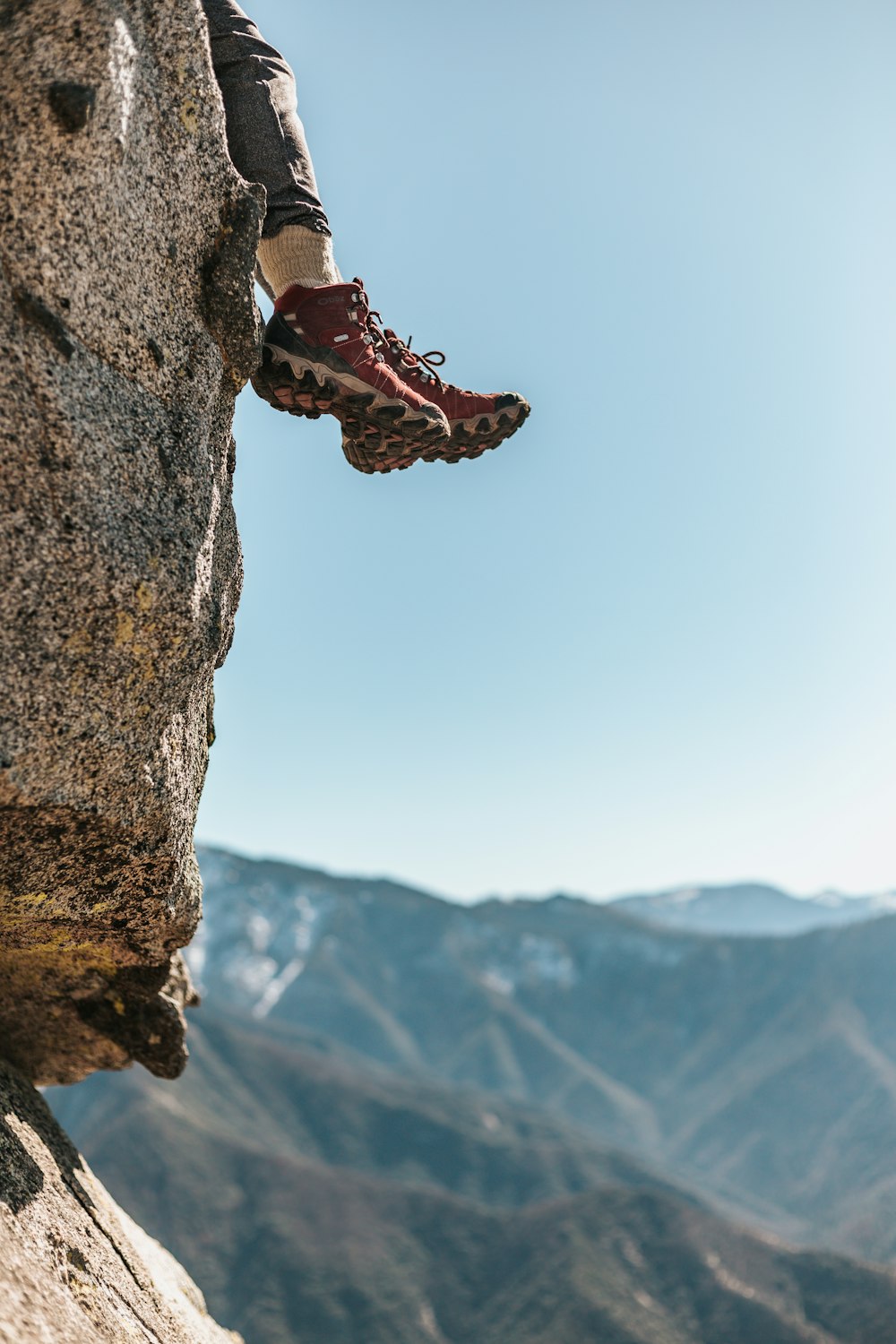 person wearing pair of red-and-white hiking shoes sitting on rocky cliff across mountains during daytime