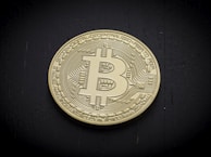 A physical representation of a Bitcoin, featuring the iconic 'B' with two vertical lines through it on the surface. The coin has intricate circuit-like patterns engraved around the edges, giving it a digital appearance. It is placed on a dark, possibly wooden surface, which provides contrast to its gold color.