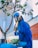 A vibrant blue parrot is perched on a gloved hand, holding a piece of food in its beak. The parrot is positioned next to a container of food and is surrounded by a small tree with green leaves. The background features a building and part of the sky.