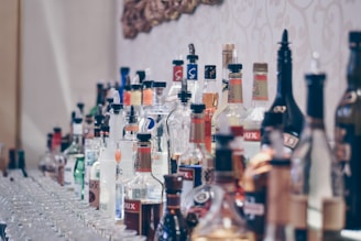 A large collection of assorted liquor bottles arranged on a shelf with labels visible. The bottles vary in shape, size, and color, with some having pour spouts attached. The background features a decorative pattern on the wall.