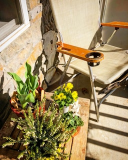 A small outdoor setup with a wooden table featuring an assortment of flower pots, including yellow flowers and green foliage, set against a stone wall. A lawn chair with a beige mesh seat and orange wooden armrests is positioned next to the table, casting shadows on the concrete floor under bright sunlight.
