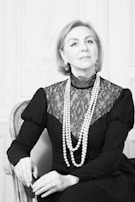 A woman is elegantly dressed in a black lace gown adorned with a long string of pearls. She sits in a vintage armchair with ornate detailing, set against an intricately designed wall. Her expression is calm and poised.