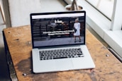A laptop is placed on a wooden table near a large window, displaying a webpage featuring an interview with a fashion and lifestyle blogger. The screen shows an outdoor urban setting with a person in the background.