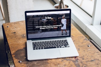 A laptop is placed on a wooden table near a large window, displaying a webpage featuring an interview with a fashion and lifestyle blogger. The screen shows an outdoor urban setting with a person in the background.