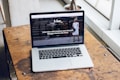 A laptop is placed on a wooden table near a large window, displaying a webpage featuring an interview with a fashion and lifestyle blogger. The screen shows an outdoor urban setting with a person in the background.