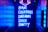 A neon sign with vibrant blue illumination displays the words 'BAR,' 'COCKTAILS,' 'DREAMS,' 'DISCO,' and 'PARTY.' Above the sign, a purple neon arrow points downward. The setting appears to be a dimly lit corridor or entrance to a venue, with walls reflecting the blue glow.