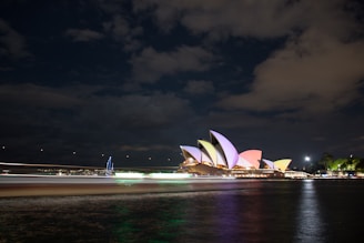 The iconic Sydney Opera House is beautifully illuminated with vibrant colors against a night sky. Light reflections shimmer on the water, creating a dynamic and lively atmosphere. Some boats' light trails appear in the foreground, adding motion to the scene.