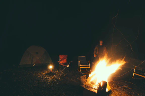 A cozy campsite at sunset with a lightweight folding chair set up beside a glowing campfire.