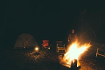 A nighttime camping scene with a brightly burning campfire in the foreground. An adult stands near the fire, surrounded by folding chairs. A pitched tent and a small lantern can be seen in the background within a wooded area.
