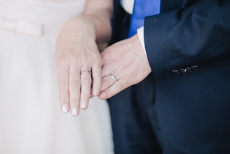 Two hands are shown overlapping. Both wear rings, suggesting a wedding or commitment ceremony. One person is dressed in a dark suit with a blue tie, while the other wears a light-colored outfit.