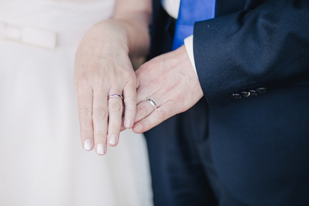 Two hands are shown overlapping. Both wear rings, suggesting a wedding or commitment ceremony. One person is dressed in a dark suit with a blue tie, while the other wears a light-colored outfit.