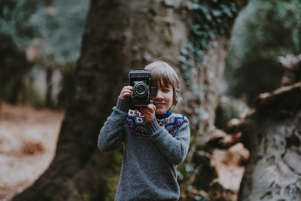 Niño sosteniendo la cámara cerca de un árbol marrón durante el día