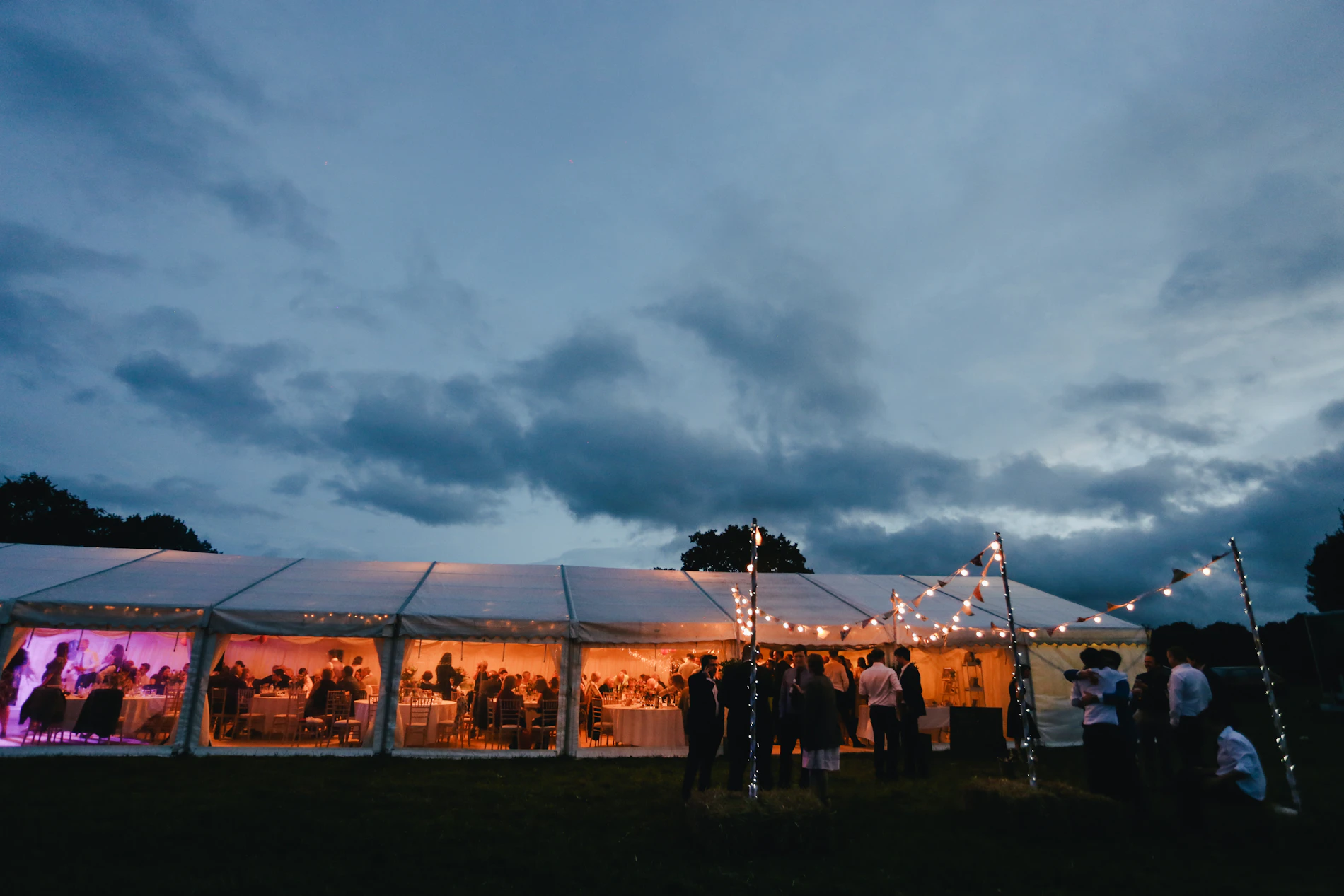 Tenda de evento iluminada por dentro ao anoitecer, com convidados em pé e céu ainda azul no horizonte