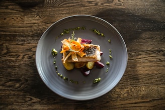 A gourmet dish featuring a piece of cooked fish garnished with thinly sliced carrots arranged on top, surrounded by a few dollops of green sauce. The fish is accompanied by roasted potatoes and beetroot cubes, all presented on a large, round, grey plate. The dish is set against a textured wooden table.