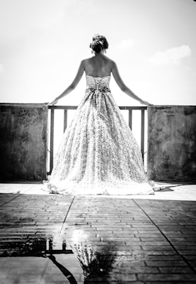 A woman in an elegant, intricately designed wedding gown stands with her back to the viewer, gazing out over a balcony. Light reflects off the floor, creating a reflection of the bride and her dress. The background is overexposed, accentuating the silhouette and creating a dreamy, ethereal feel.