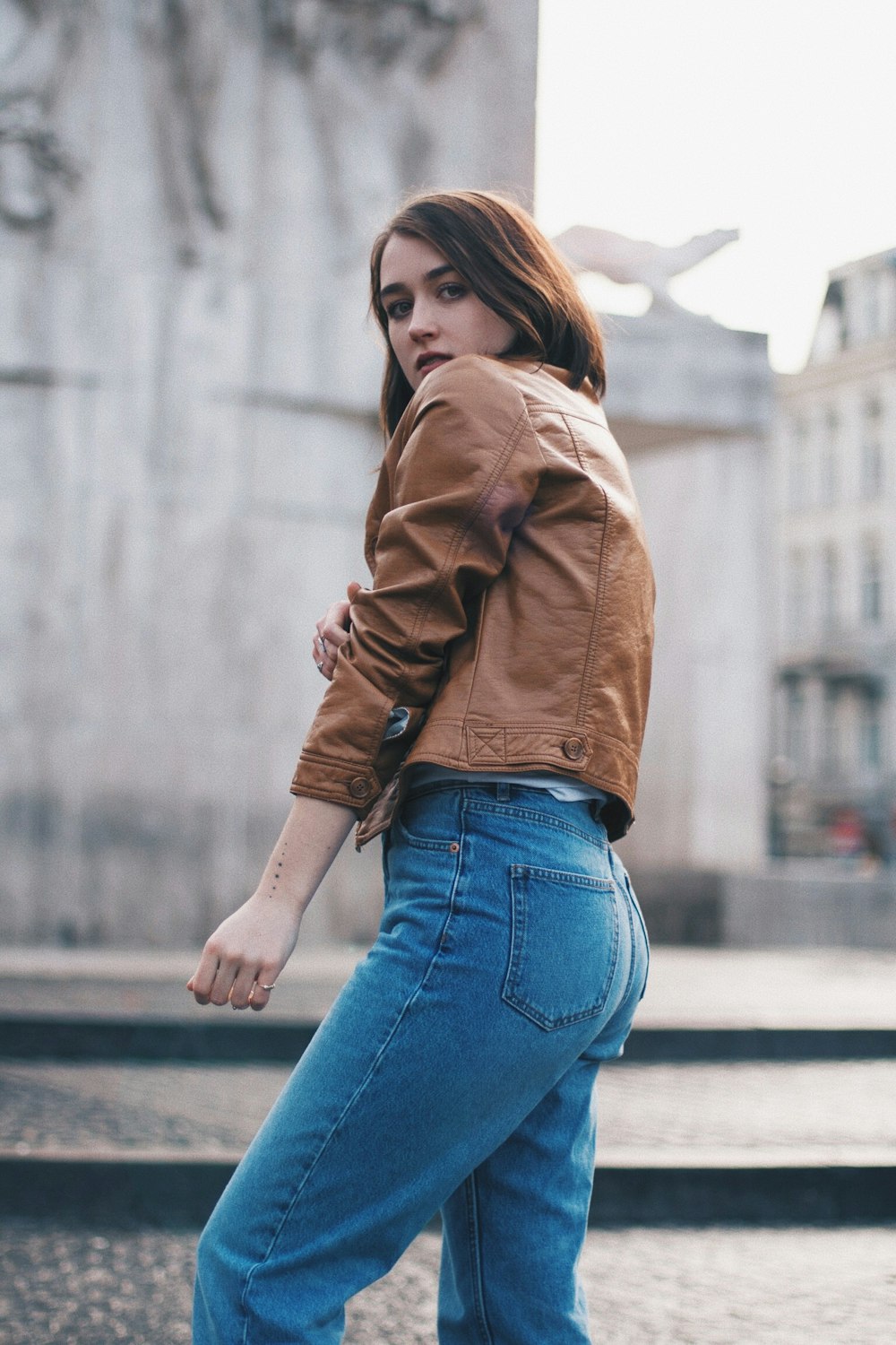 selective focus photography of woman in brown leather jacket stands near concrete building